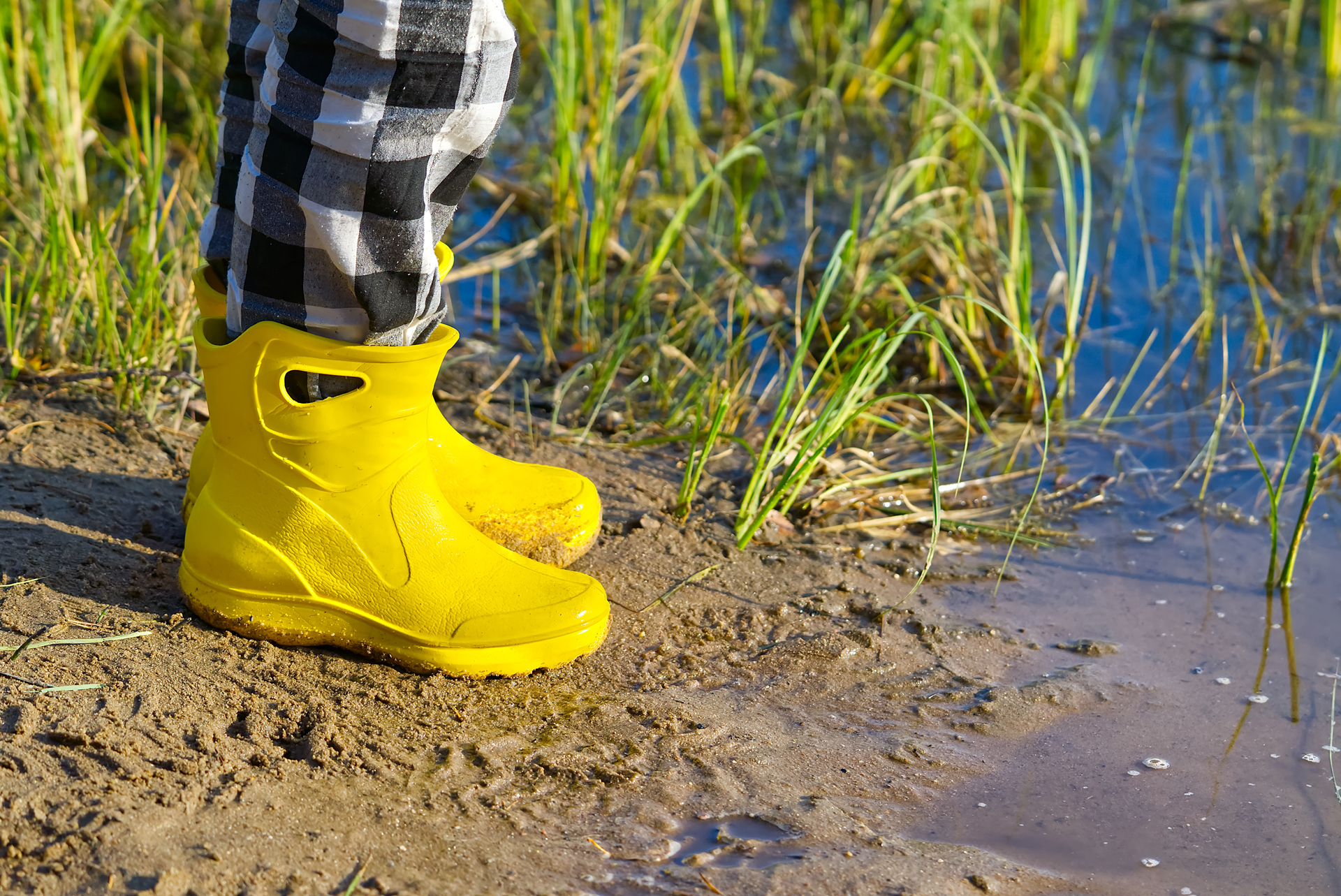 A person is standing in shallow water, wearing bright yellow rubber boots. The water is clear, revealing the sandy bottom and occasional patches of grass, creating a picturesque beach scene. A person is standing in shallow water, wearing bright yellow rubber boots. The water is clear, revealing the sandy bottom and occasional patches of grass, creating a picturesque beach scene.